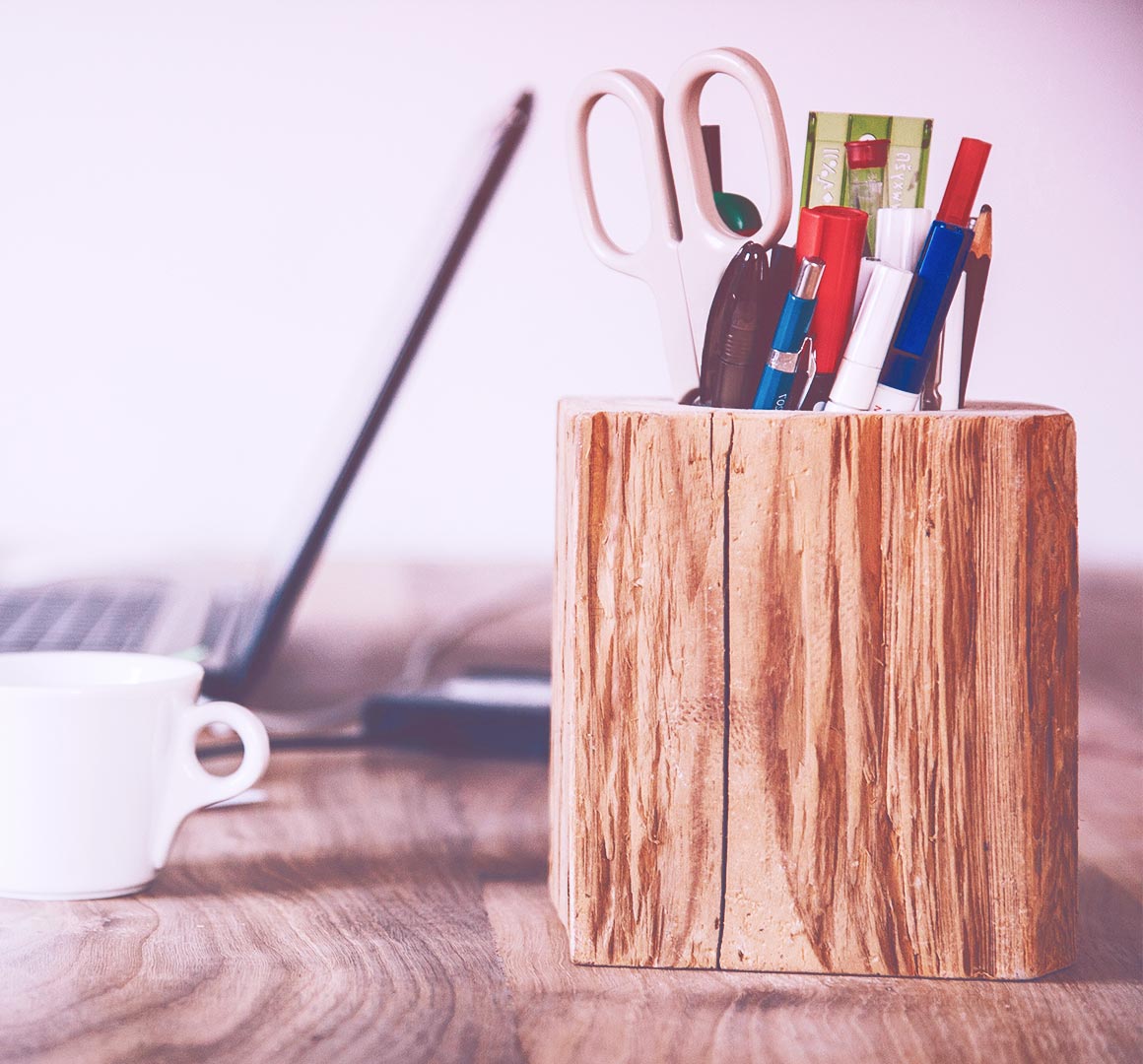 A wooden pencil/pen holder setting on a desk with a laptop and coffee cup in the background